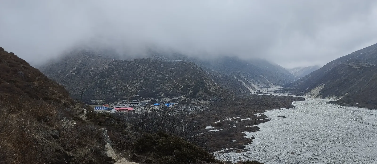 First sight of Khambachen's lodges at the head of a wide gravel riverbed under low cloud