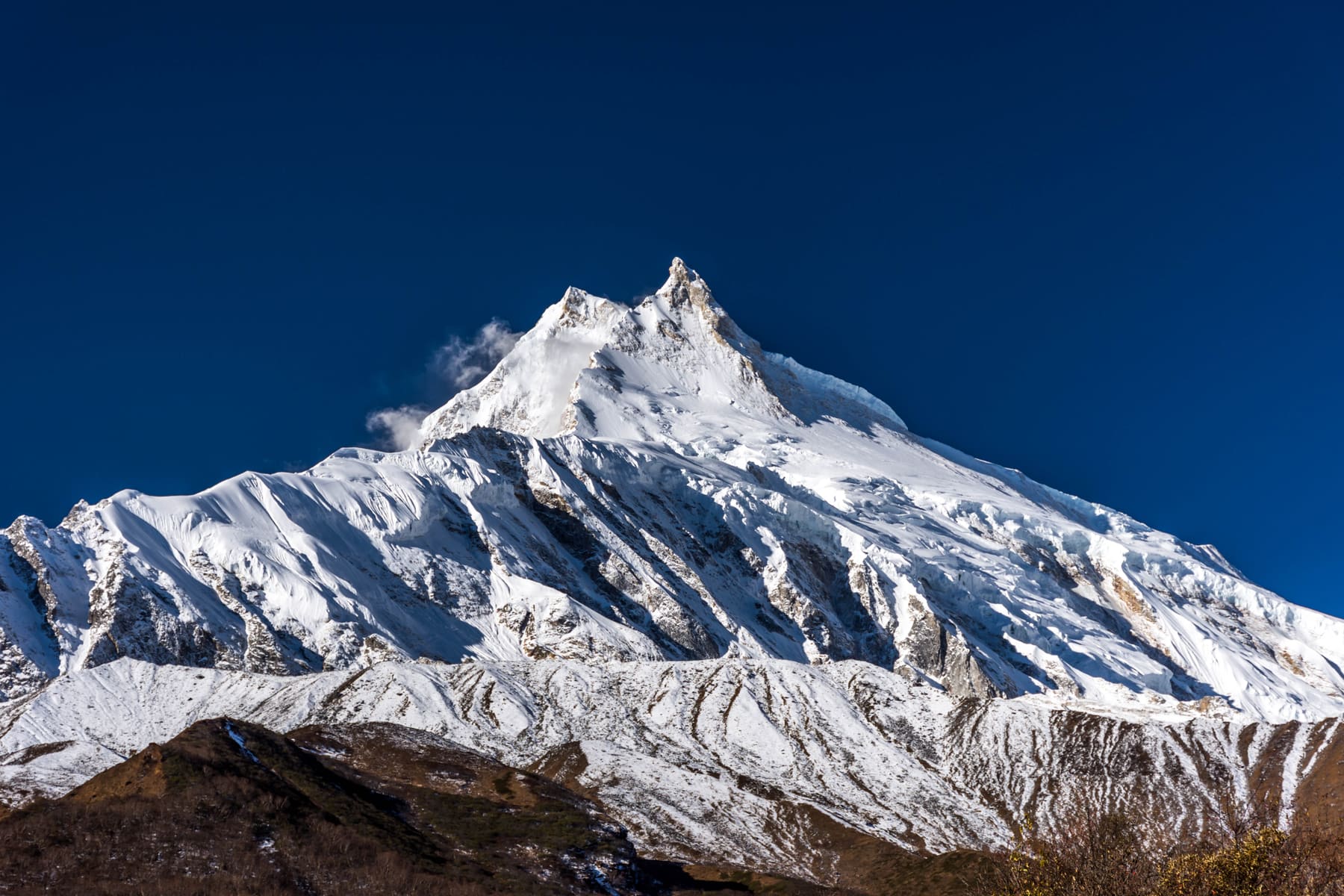 Manaslu's summit ridge (8,163m), snow plume trailing off the peak, seen from the approach trail.