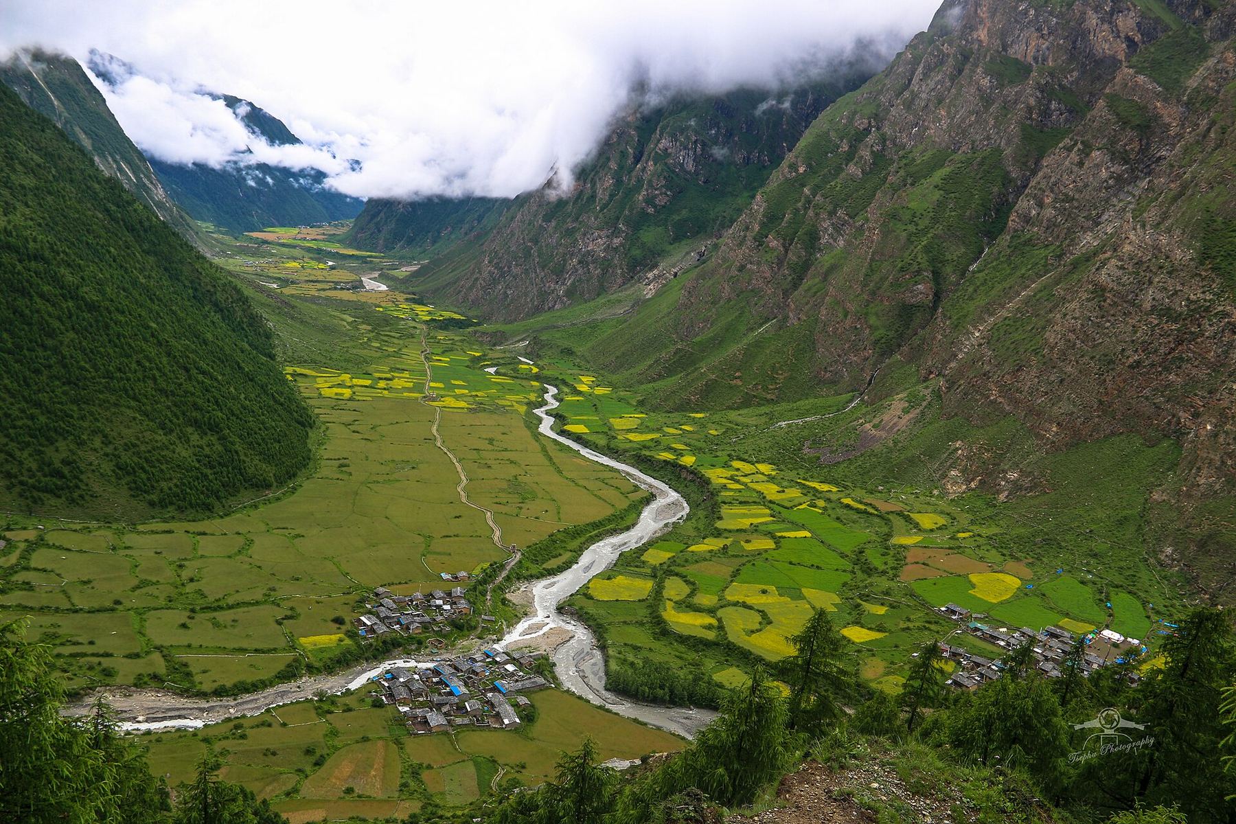 The Tsum Valley from above: a river winding through terraced fields and villages between steep forested and rocky slopes, low cloud overhead.
