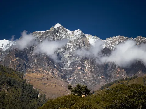 A rocky snow peak with cloud drifting across its face above forested slopes