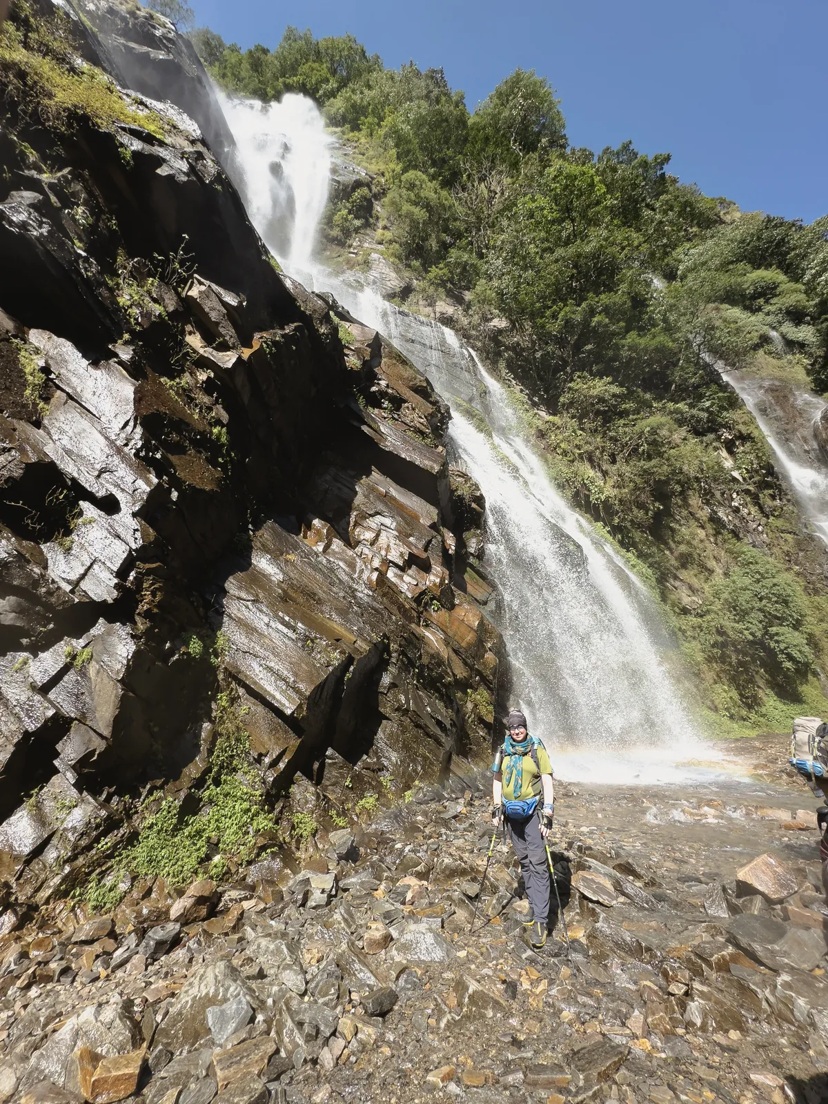 A trekker on a rocky trail at the foot of a waterfall pouring off a cliff